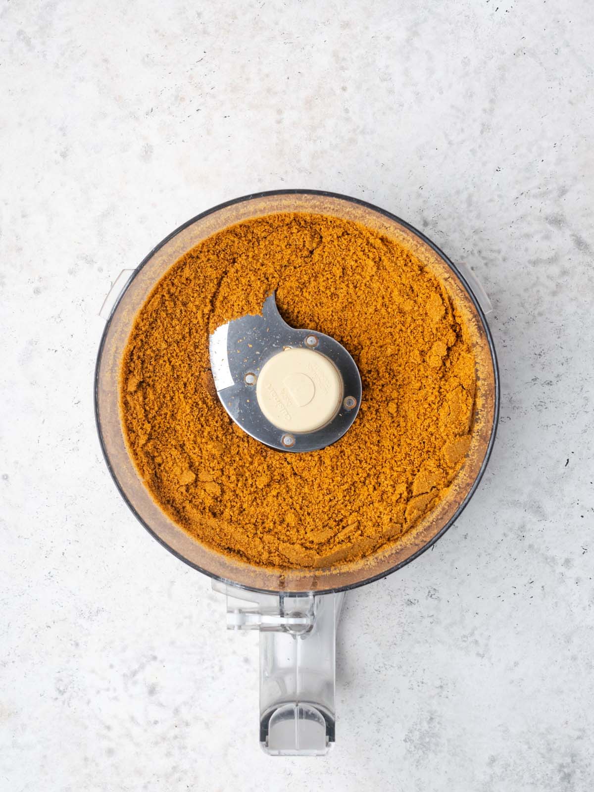 A food processor bowl filled with finely crushed golden-brown cookie crumbs and a metal blade.