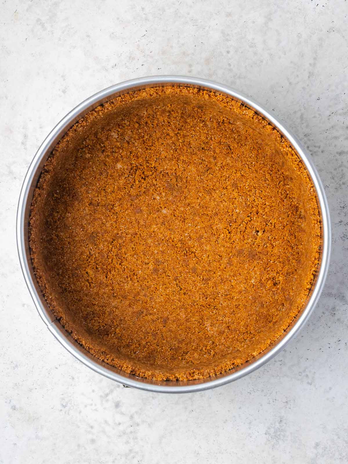 A circular springform pan containing a pressed graham cracker crust.
