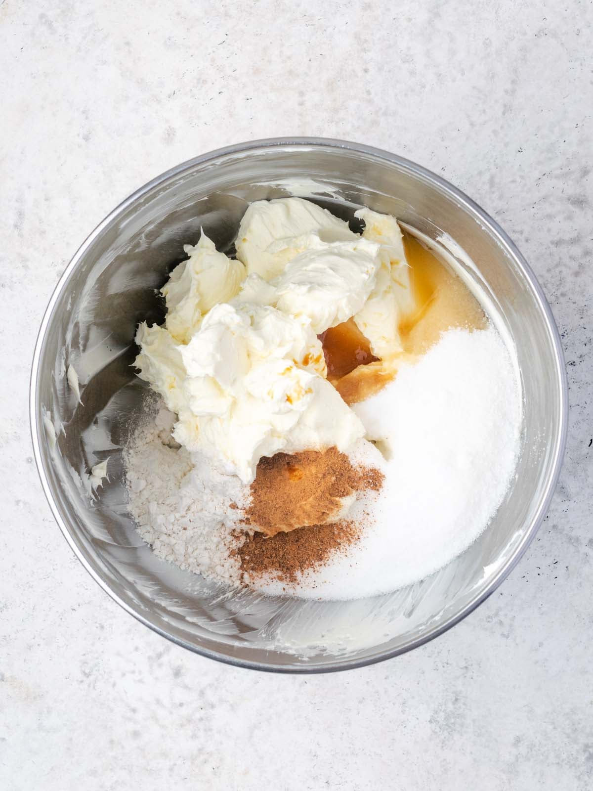 A metal mixing bowl containing cream cheese, sugar, flour, cinnamon, and liquid ingredients.
