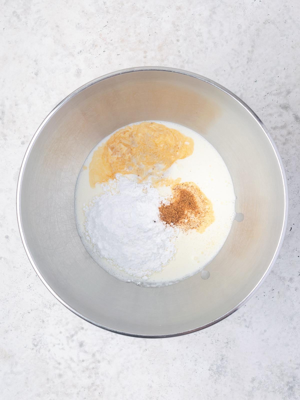 A metal mixing bowl containing milk, powdered sugar, a golden liquid, and a dash of cinnamon.