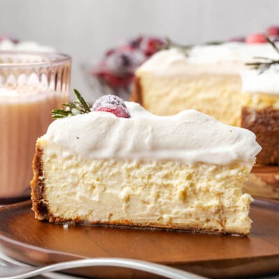A slice of pay de queso topped with whipped cream, a sugared cranberry, and a rosemary sprig on a wooden plate.
