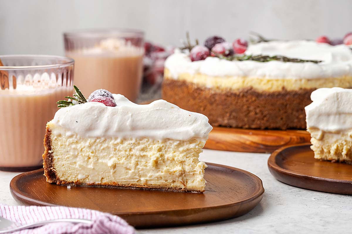 A slice of pay de queso topped with whipped cream, a sugared cranberry, and a sprig of rosemary on a wooden plate, with a whole cheesecake in the background.