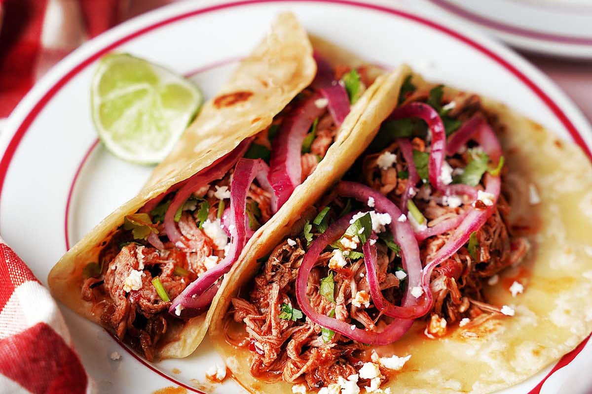 Two soft tacos filled with shredded meat, topped with pickled red onions, cilantro, and crumbled cheese, served with a lime wedge on a plate.